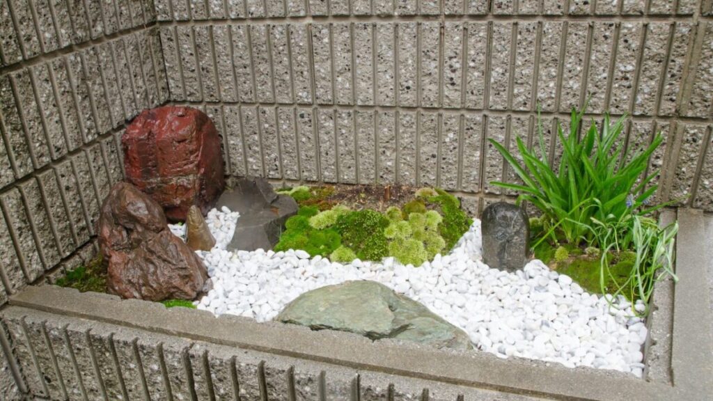 玄関横の小さなスペースに白い玉砂利と庭石を下草、苔がある和風スペースの写真