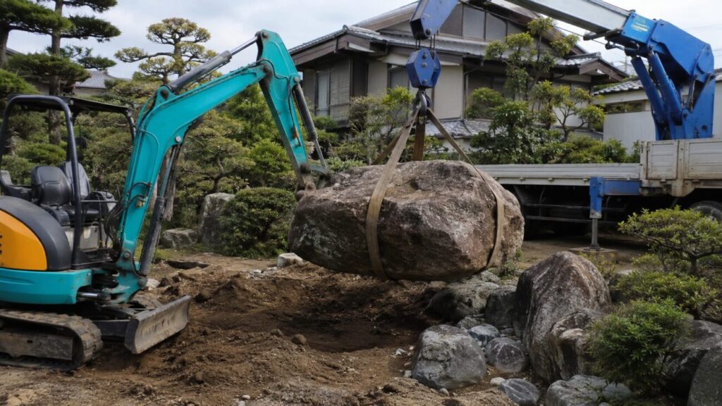 重機とクレーンで大きな庭石を吊り上げている撤去作業の様子。庭石撤去費用が高くなるケースを説明する画像