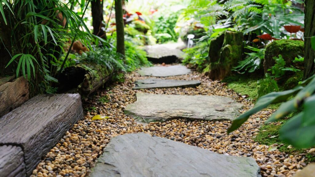 鉄平石の飛び石の周りに茶色の砂利が敷かれており、苔や下草がある自然な和風庭園の写真